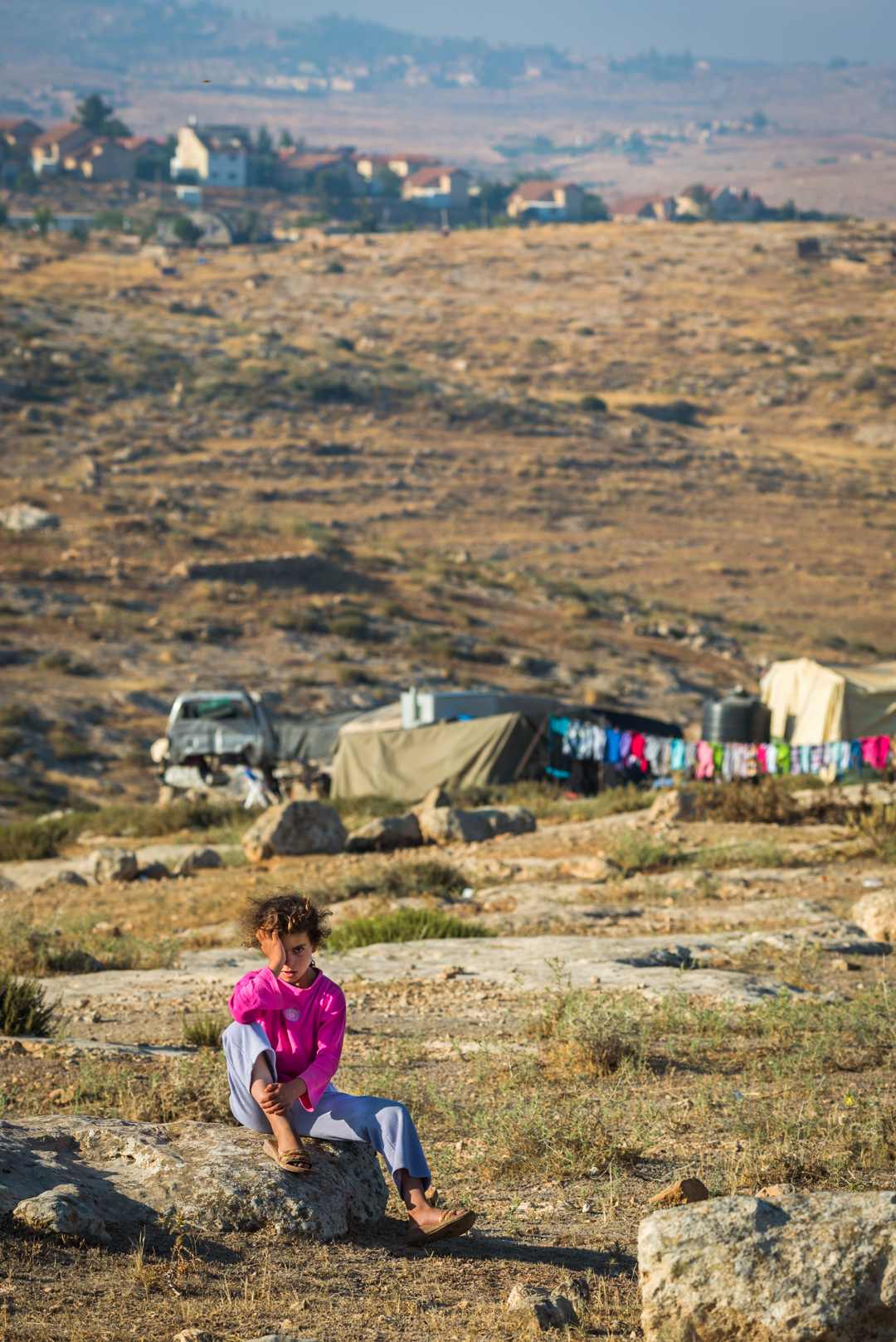 A Palestinian girl sits in front of Susiya, her village, overlooked by an illegal Israeli settlement on the hill. Although she's a child, she understands that because of the settlement, the Israeli authorities will soon demolish her home and village.