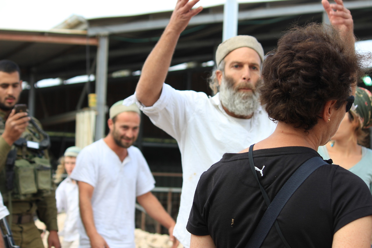 A settler yells and gestures at a peace activist, while in the background an Israeli soldier films the scene on his cell phone.